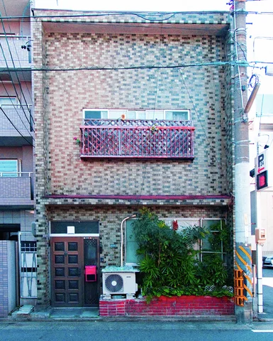 A narrow patterned-brick house has a pink window railing and plants in a red planter.