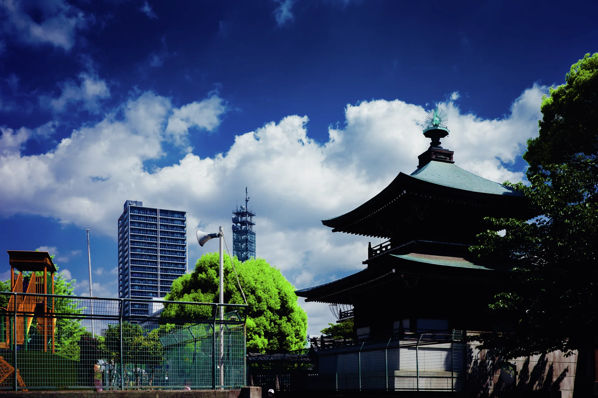 A dark tiered-roof building stands beside a fenced playground and modern towers beneath bright clouds.