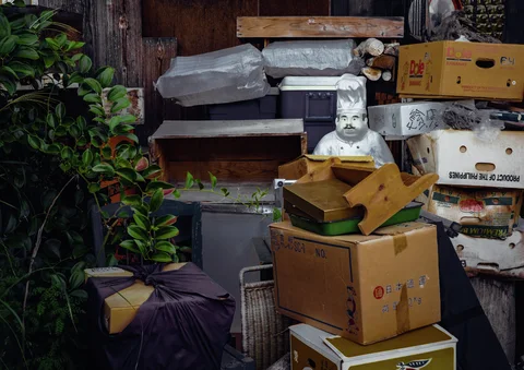A small chef figure stands among stacked cardboard boxes, containers and miscellaneous objects beside foliage.