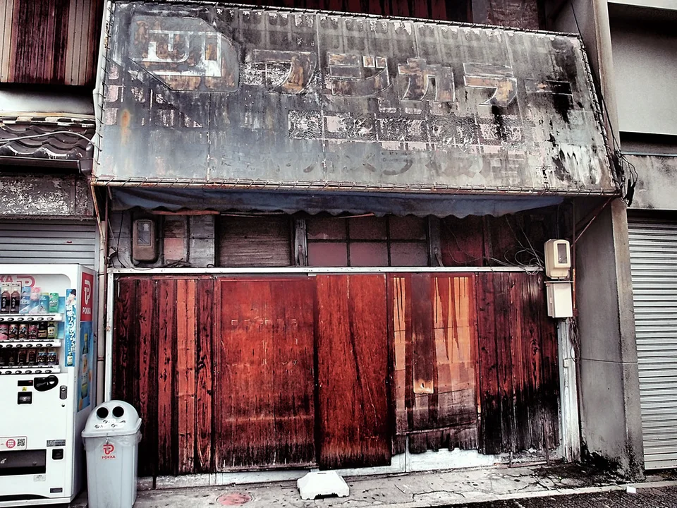 A severely faded sign tops reddish wooden storefront doors beside a vending machine and bin.