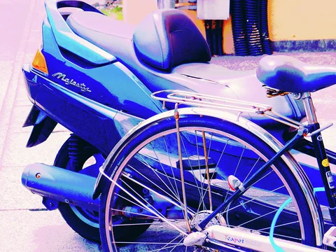 A bicycle wheel and rear rack overlap a blue scooter with a tall padded seat.