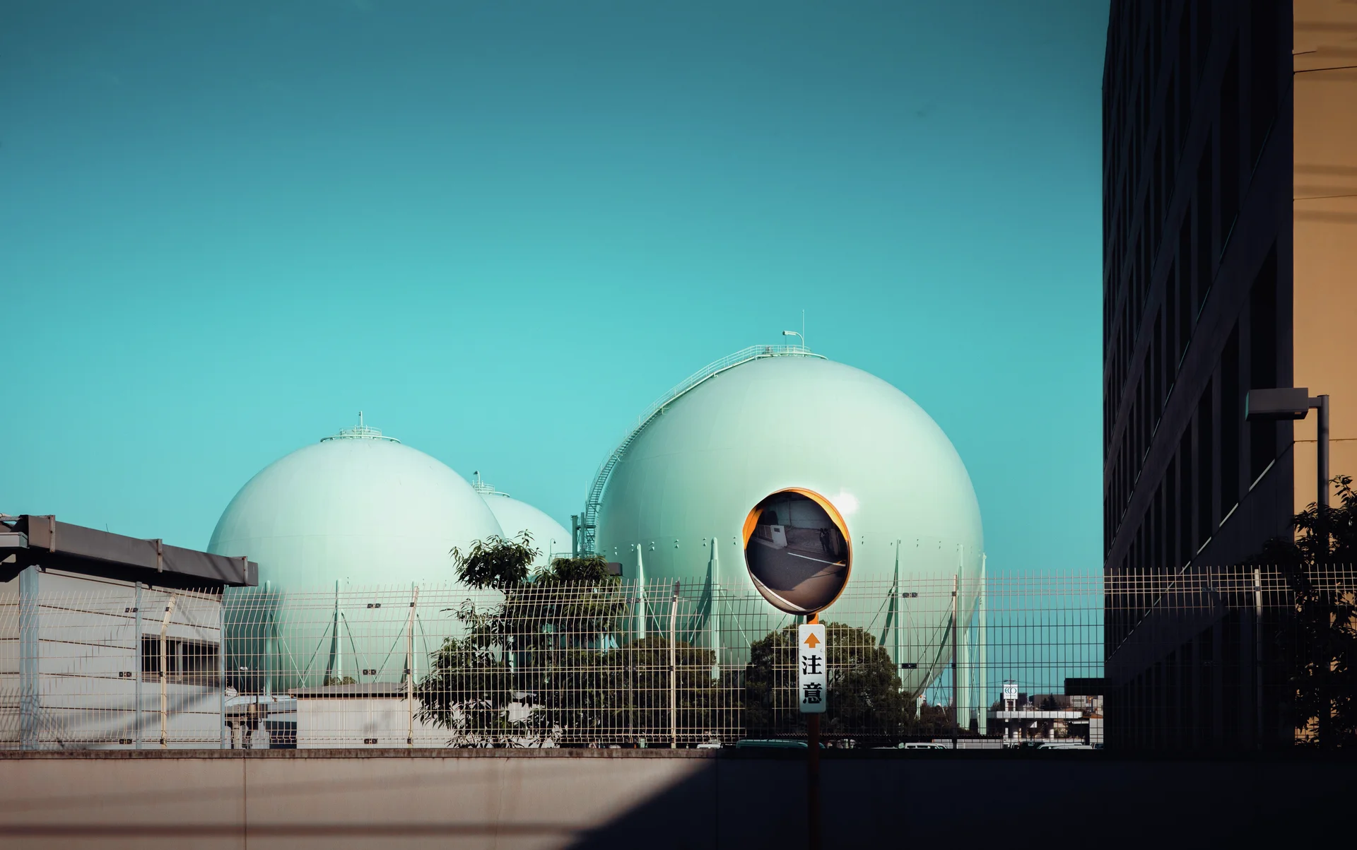 Two pale green spherical industrial tanks stand behind a fence beneath turquoise sky beside a roadside mirror.