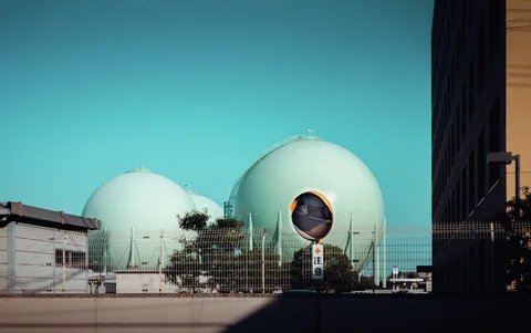 Two pale green spherical industrial tanks stand behind a fence beneath turquoise sky beside a roadside mirror.