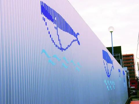 Blue stylized fish and pale wave marks decorate a long white corrugated wall beside a narrow walkway.