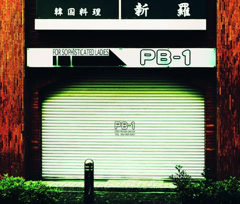 A closed pale shutter marked PB-1 sits beneath dark shop signs and warm brick walls.