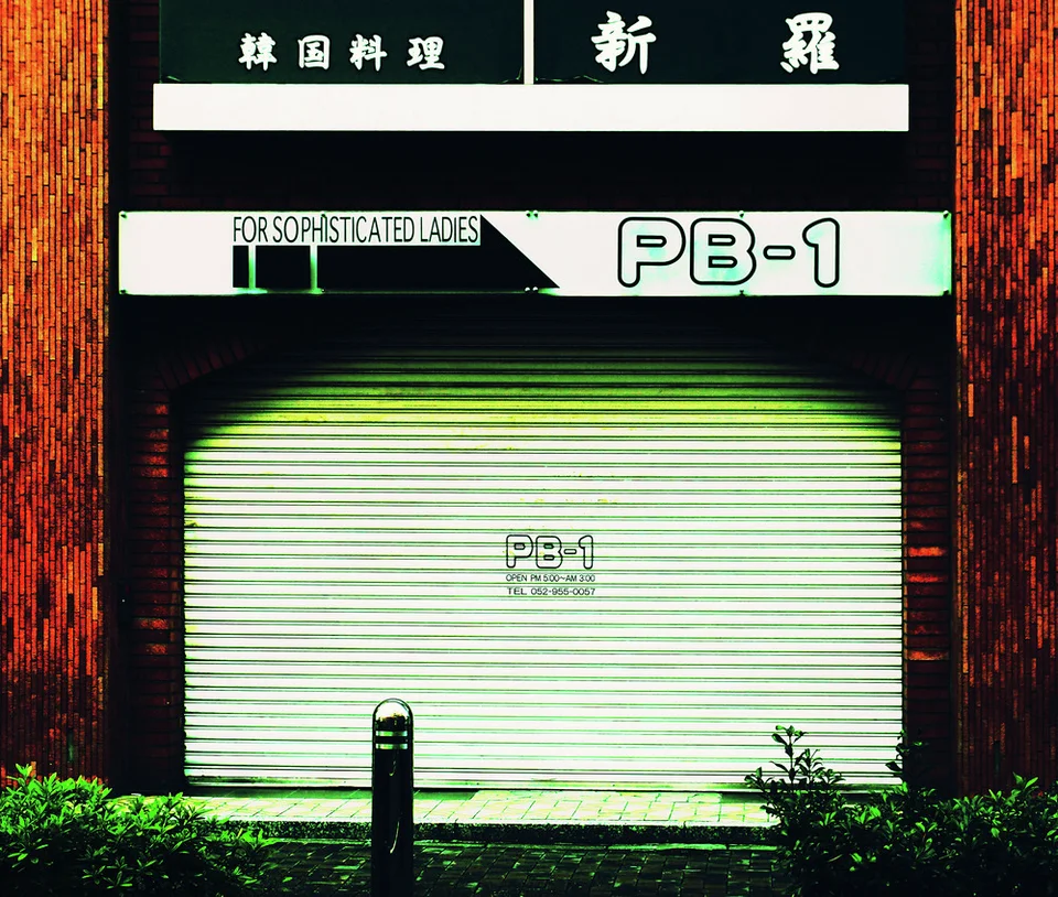 A closed pale shutter marked PB-1 sits beneath dark shop signs and warm brick walls.